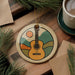 A round ornament featuring a guitar design is placed on a wooden surface, surrounded by pine branches and a cup of coffee.