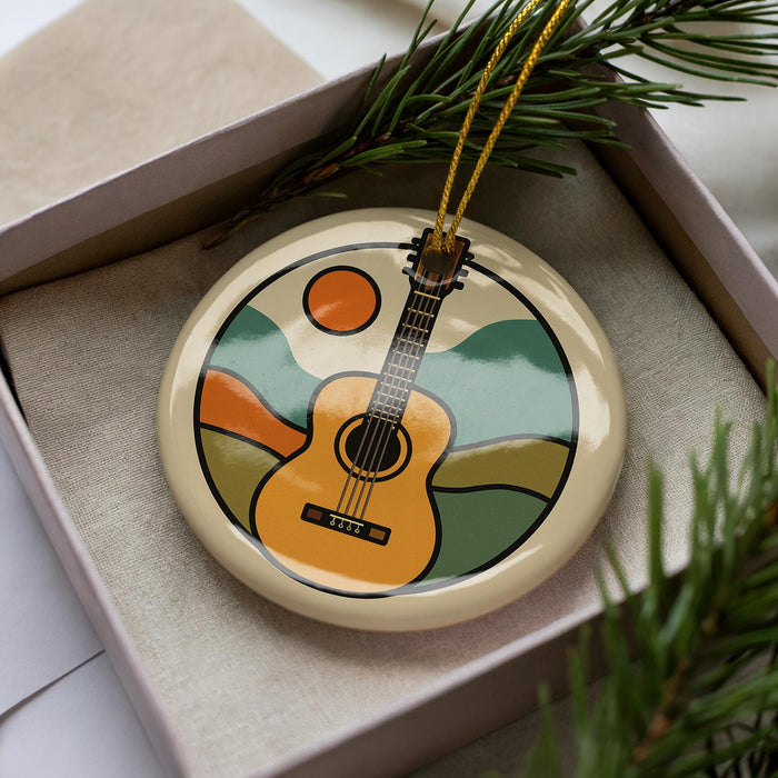 A round ornament with a guitar design is displayed in a box, surrounded by pine branches.