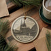 A decorative ornament featuring the Cologne Cathedral in Germany, with a cup of coffee and pine branches in the background.