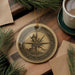 A decorative compass ornament with a lighthouse on a wooden surface, surrounded by pine branches and a cup of coffee.