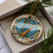 A decorative ornament featuring a harp is displayed in a box, surrounded by pine branches.