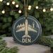 A circular ornament with a graphic of an airplane and a mountain range, along with the word "DEN" written on it, is displayed in front of a blurred background with Christmas lights.