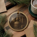 A round ornament featuring an image of a distillery or brewing equipment, such as a copper still or kettle, is displayed on a wooden surface alongside a cup of coffee and some pine branches.