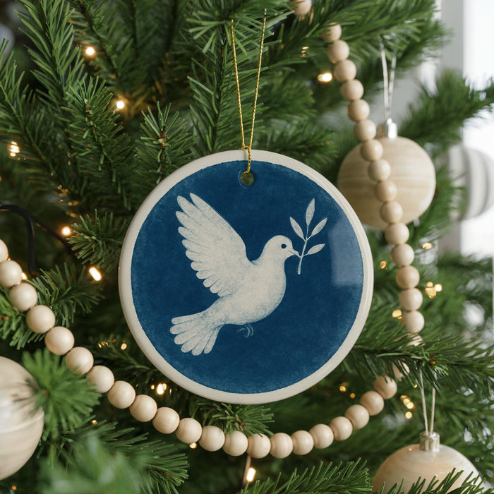 A blue and white ceramic ornament featuring a white dove in flight is hanging on a Christmas tree, surrounded by white beads and greenery.