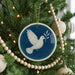 A blue and white ceramic ornament featuring a white dove in flight is hanging on a Christmas tree, surrounded by white beads and greenery.