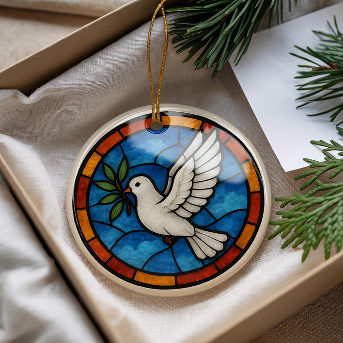 A stained glass ornament in the shape of a white dove with outstretched wings is displayed in a box, surrounded by pine branches and a white card.