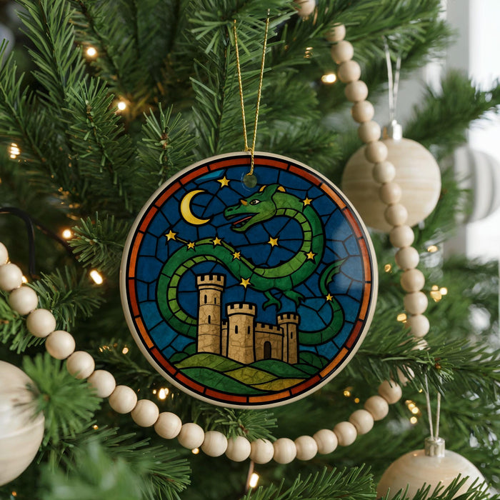 A stained glass ornament with a castle and a dragon is hanging on a Christmas tree, surrounded by ornaments and lights.