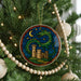 A stained glass ornament with a castle and a dragon is hanging on a Christmas tree, surrounded by ornaments and lights.