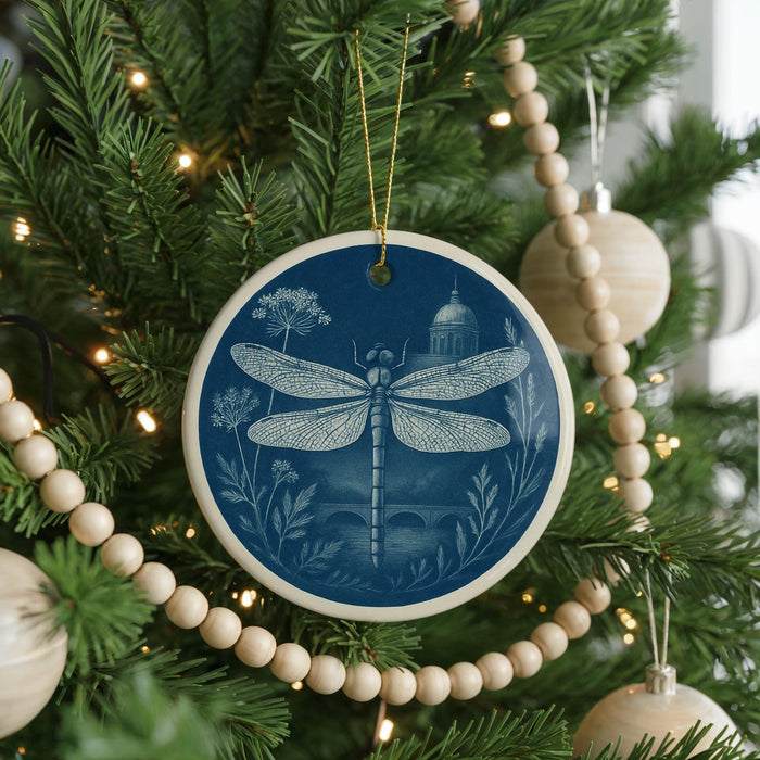 A blue ceramic ornament with a dragonfly design hangs from a Christmas tree, surrounded by white beads and greenery.