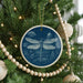 A blue ceramic ornament with a dragonfly design hangs from a Christmas tree, surrounded by white beads and greenery.
