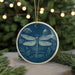 A blue ceramic ornament featuring a detailed illustration of a dragonfly is displayed on a table, surrounded by pine branches and twinkling lights.