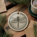 a ceramic ornament with a detailed illustration of a dragonfly, placed on a wooden surface alongside a cup of coffee and some pine branches.