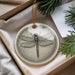 A ceramic ornament in the shape of a dragonfly, with a detailed illustration of the insect, is displayed in a box with pine branches.