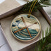 A circular ornament with a design featuring a pair of drumsticks crossed over each other, set against a backdrop of a wave and mountains.