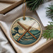 A circular ornament with a design featuring a wave and a mountain, with two drumsticks crossed in the middle, is displayed in a box with pine branches.