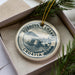 A decorative ornament in a box, featuring a circular design with text and an illustration of a coastal town.