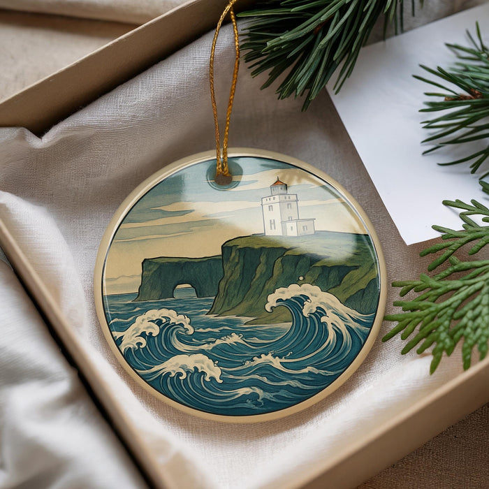 A round ornament with a lighthouse on a cliff, surrounded by waves crashing against the shore, is displayed in a box with pine branches.