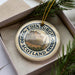A decorative ornament featuring the image of Edinburgh Castle, a famous landmark in Scotland, is displayed in a box with pine branches.