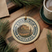 A decorative ornament featuring the Edinburgh Castle in Scotland, with the text "EDINBURGH SCOTLAND" prominently displayed. The ornament is placed on a wooden surface, surrounded by pine branches and a cup of coffee.
