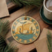 a decorative ornament with a sailboat and a person on a wooden surface, surrounded by pine branches and a cup of coffee.