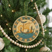 A round ornament with a sailboat and a person on a boat is hanging on a Christmas tree, surrounded by ornaments and lights.