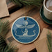 a blue ceramic ornament with a design depicting a person in a boat on a starry night, surrounded by pine branches and a cup of coffee.