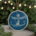 A blue circular ornament with a white illustration of a person in a boat on a starry night, surrounded by a starry sky.
