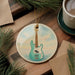 A ceramic ornament featuring a blue bass guitar is displayed on a wooden surface, surrounded by pine branches and a cup of coffee.