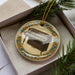 A decorative ornament featuring a piano is displayed in a box, surrounded by pine branches.