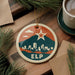 A circular ornament with a cityscape design and a star, featuring the text "ELP" on it, is displayed on a wooden surface alongside a cup of coffee and some pine branches.