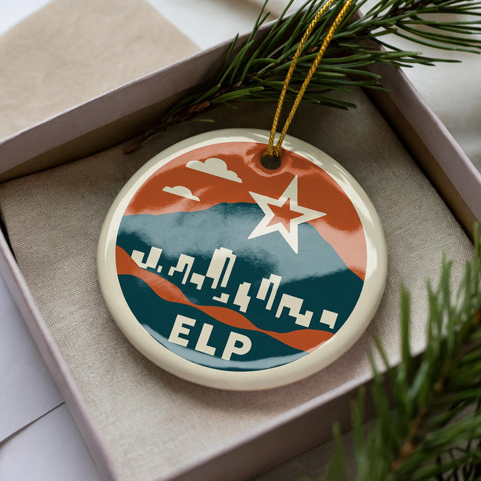 A circular ornament with a city skyline design and a star, placed in a box with pine branches.