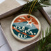 A circular ornament with a city skyline design and a star, placed in a box with pine branches.