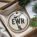 A circular ornament with the letters "EWR" and an image of the Statue of Liberty is displayed in a box with pine branches.