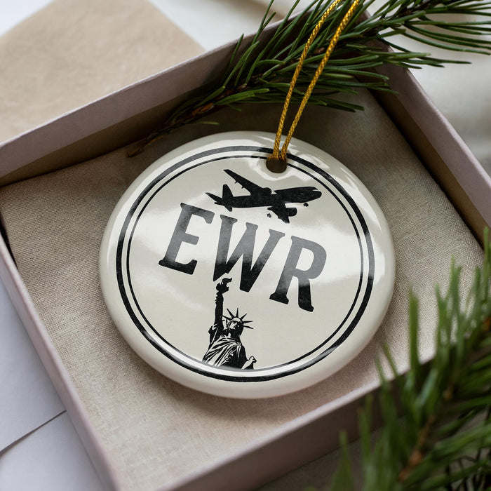 A white ceramic ornament with the letters "EWR" and a silhouette of the Statue of Liberty, placed in a box with pine branches.