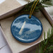 A blue ornament with a lighthouse and a house is displayed in a box, surrounded by pine branches.