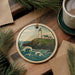 A round ornament featuring a lighthouse on a cliff overlooking the ocean, surrounded by pine branches and a cup of coffee on a wooden surface.