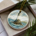 A round ornament featuring a lighthouse on a rocky island, surrounded by water and clouds, is displayed in a box with pine branches.