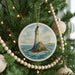 A round ornament featuring a lighthouse on a rocky island, surrounded by a blue ocean and white clouds, is hanging on a Christmas tree.