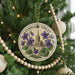 A round ornament on a Christmas tree, featuring a floral design with purple flowers and a church spire.