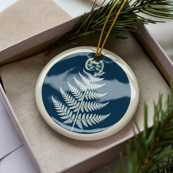 A blue and white ceramic ornament with a white fern design is displayed in a box, surrounded by pine branches.