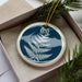 A blue and white ceramic ornament with a white fern design is displayed in a box, surrounded by pine branches.
