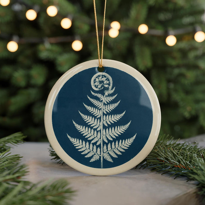 A blue and white circular ornament featuring a fern design is displayed in the center of the image, surrounded by pine branches and twinkling lights in the background.