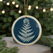 A blue and white circular ornament featuring a fern design is displayed in the center of the image, surrounded by pine branches and twinkling lights in the background.