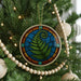 A Christmas tree decorated with a colorful stained glass ornament featuring a fern design, surrounded by wooden beads and other ornaments.