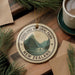 A circular ornament with the text "FIORLAND NATIONAL PARK NEW ZEALAND" and a mountain landscape design, hanging on a wooden surface.