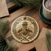 A round ornament with a design featuring a fire lookout tower, crossed axes, and a laurel wreath, placed on a wooden surface next to a cup of coffee and some pine branches.