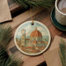 A decorative ornament featuring a painting of the Florence Cathedral, also known as the Cathedral of Saint Mary of the Flower, is displayed on a wooden surface alongside a cup of coffee and some pine branches.