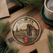 A circular ornament with a castle and a river, featuring the text "GDASK" in the center, is displayed on a wooden surface alongside a cup of coffee and some pine branches.