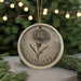 A decorative ornament featuring a detailed illustration of a flower, with the word "JANUARY" written below it, is displayed on a table with pine branches in the background.