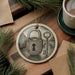A ceramic ornament featuring a lock and key design, surrounded by pine branches and a cup of coffee on a wooden surface.